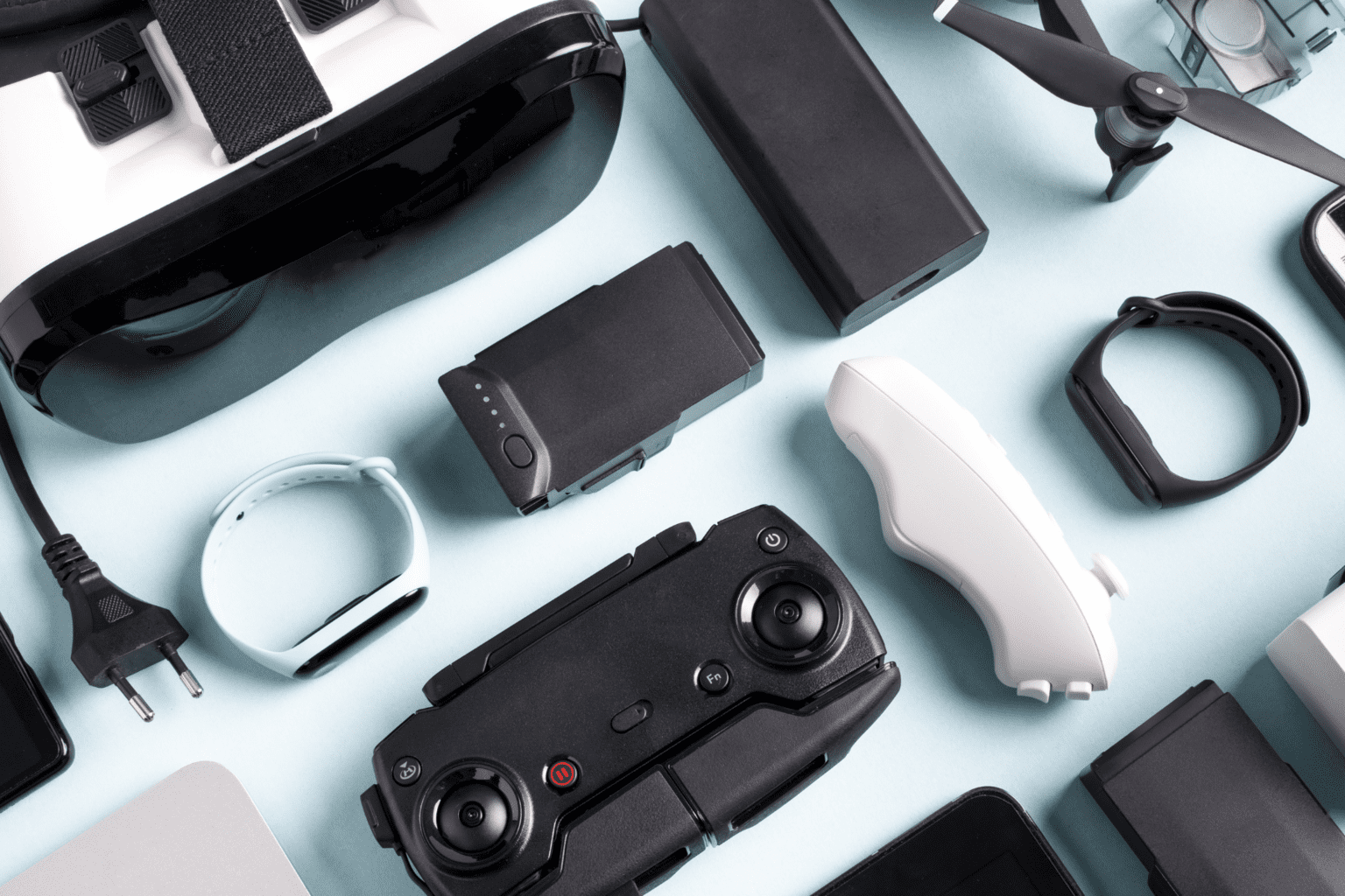 A flat lay of modern educational and tech gadgets including a VR headset, smartwatch, drone, remote controller, power bank, and wearable devices on a light blue background.